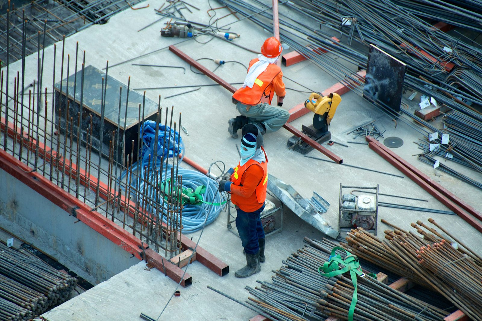 Two workers on a concrete deck laid with reinforcing bar — one kneeling at a cut-off saw, one standing over a coiled hose beside bundled steel