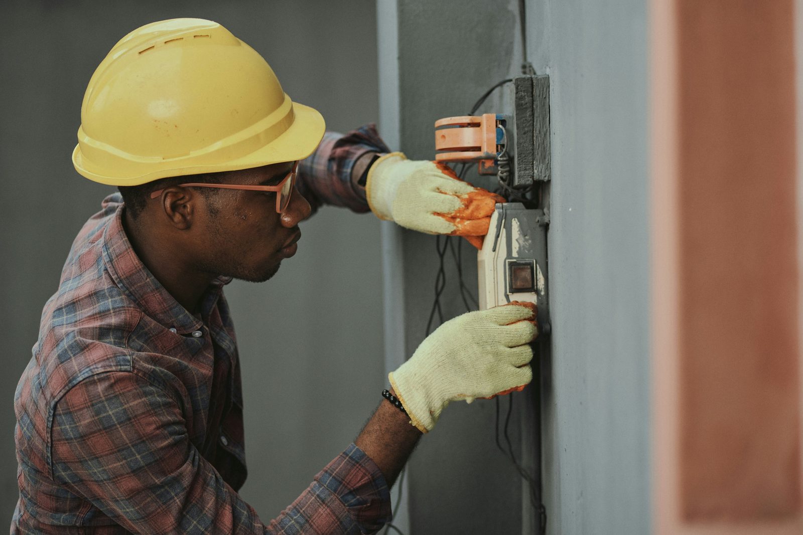 An electrician in a hard hat and work gloves wiring a device into an enclosure on a concrete wall