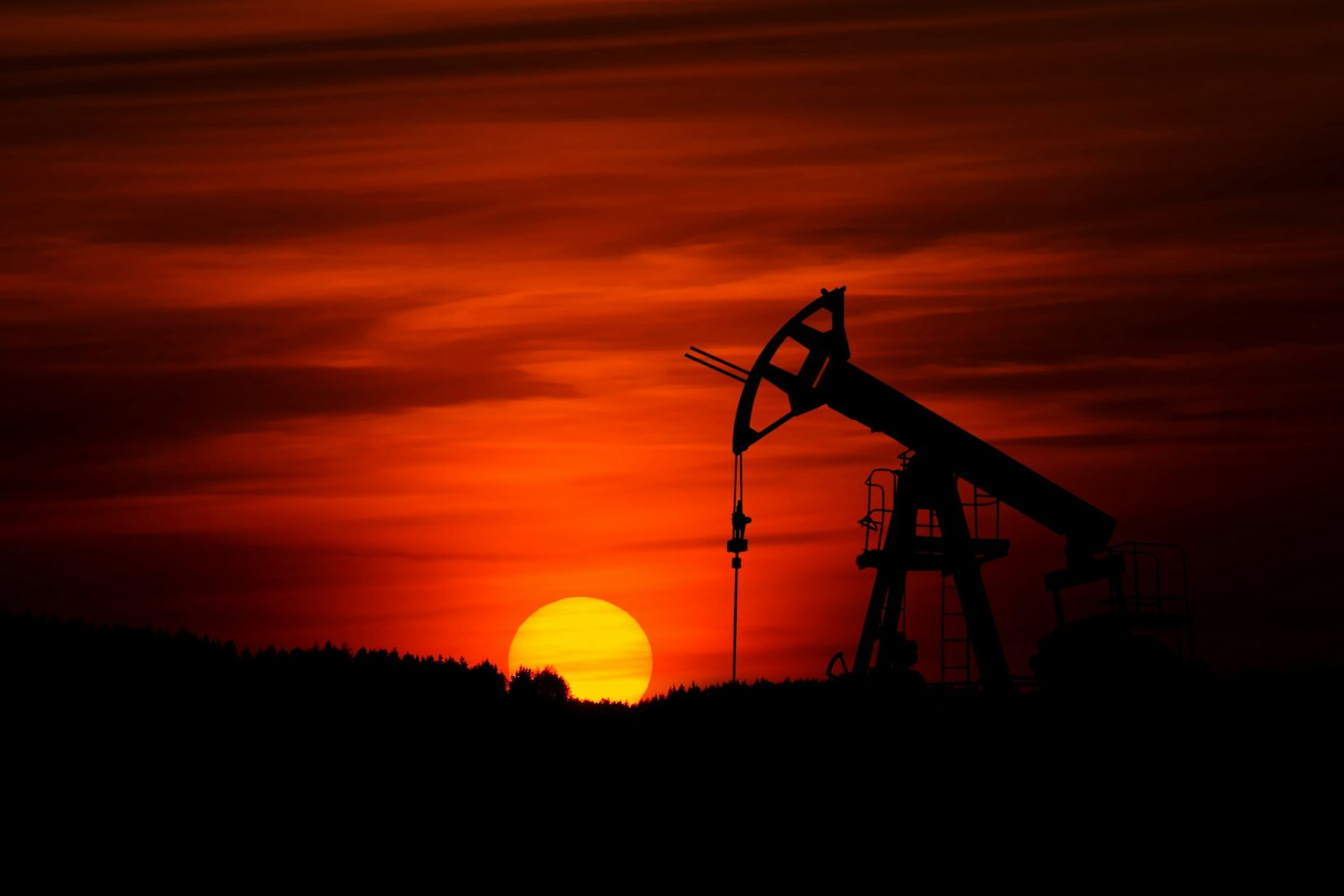 A pumpjack silhouetted against a deep red sunset over a distant treeline
