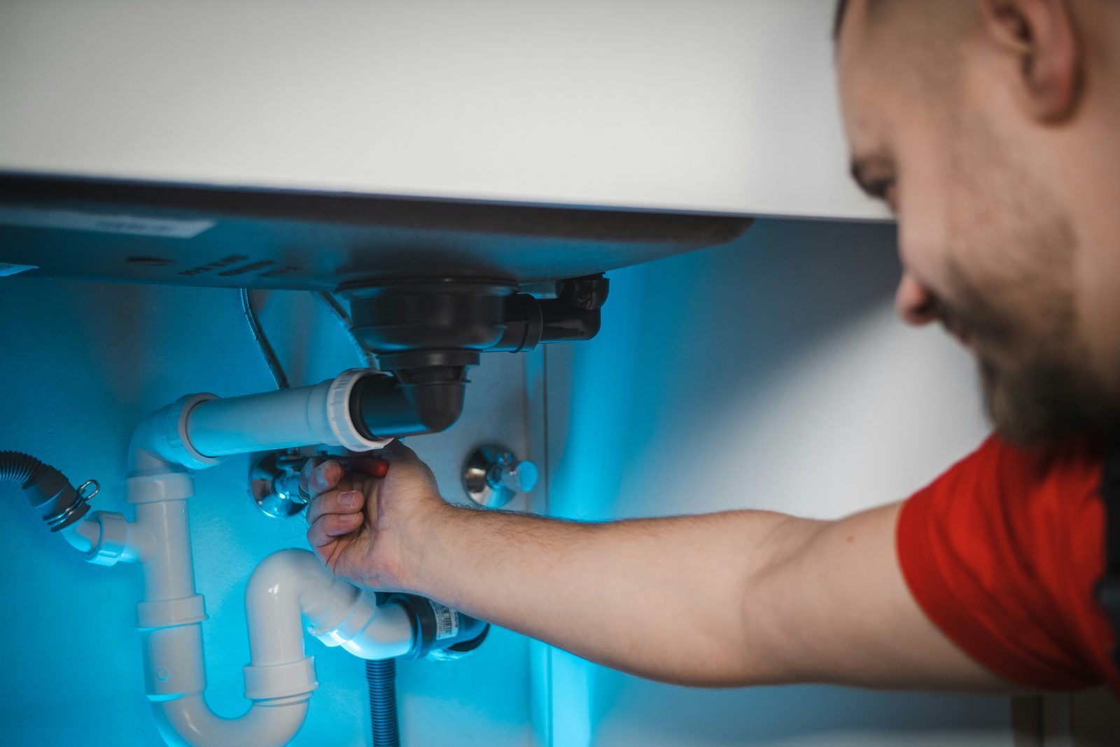 A plumber reaching into the cabinet under a sink with both hands on a white plastic P-trap, the enclosed space lit blue