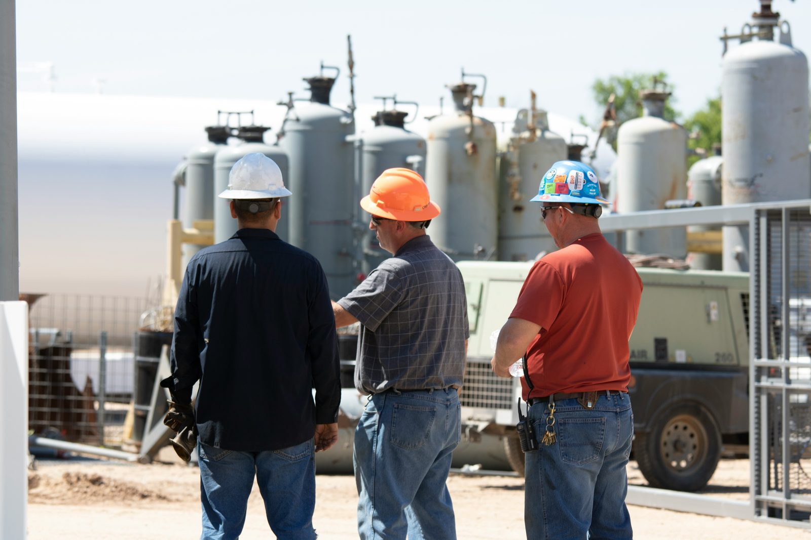 Three workers in hard hats standing together at a tank battery, mid-conversation, one gesturing towards the vessels