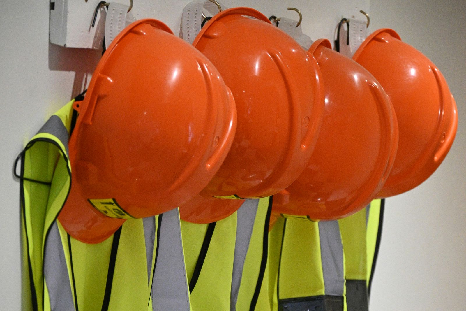 Four sets of orange hard hats and hi-vis vests hanging together on a row of hooks, indoors, before a shift
